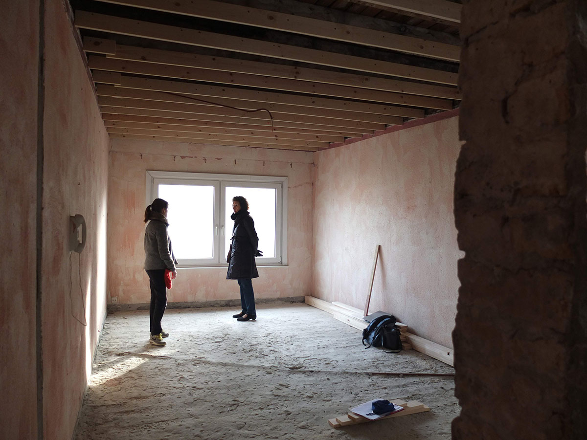 en cours de chantier : vue depuis le hall vers le salon-cuisine, avec architecte et client en cours de discussion au milieu de la pièce vide, murs sols et plafonds à nu